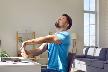Working man takes a break for stretching exercise. Man in blue T shirt sitting at desk with laptop computer at home and doing relaxing moves to prevent muscle pain and discomfort in arms and shoulders - obrazy, fototapety, plakaty