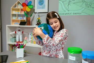 Lovely female student with closed eyes hugging with her hands a handmade globe world at ecology classroom. Sustainable lifestyle education concept.
