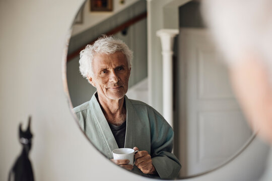 Smiling senior man with coffee cup looking at himself in mirror at home
