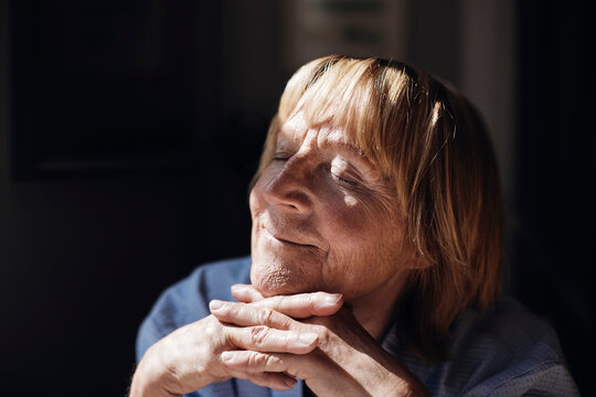 Smiling blond senior woman with hands on chin relaxing in sunlight at home