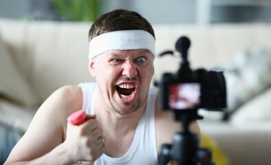 Portrait of an emotionally male blogger with grimace on face and holding dumbbells in front of camera. Sloth fatigue and daily sports training - obrazy, fototapety, plakaty