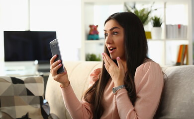 Woman surprised in shock looks at smartphone screen while sitting on couch at home. Emotion joy delight and good news concept - obrazy, fototapety, plakaty