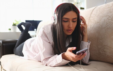 Young woman in headphones is relaxing on sofa and listening to music online using smartphone. Music and educational apps for phone