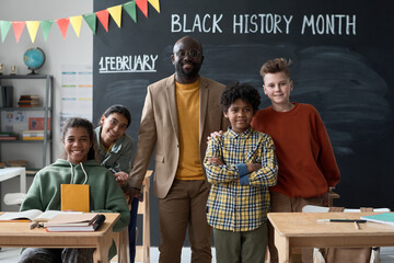 Portrait of African AMerican teacher smiling at camera together with his pupils while they standing in classroom against blackboard