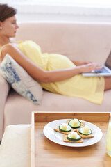 Obraz na płótnie Canvas Pregnant woman, food and laptop on sofa with healthy, nutrition or wellness for lunch or snack in living room. Person, meal or brunch and crackers on couch for remote work, relax and internet at home