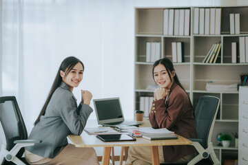 Fototapeta na wymiar Two cute Asian businesswomen in suits talking in modern workplace. Thai woman. Southeast Asian woman. looking at laptop together in office