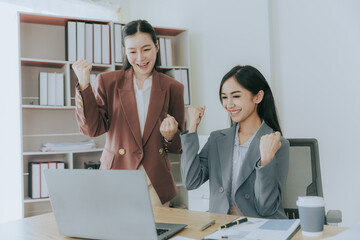 Two cute Asian businesswomen in suits talking in modern workplace. Thai woman. Southeast Asian woman. looking at laptop together in office