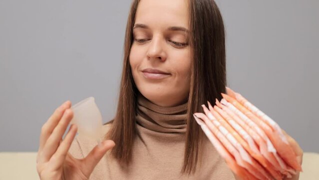Smiling brown haired woman holding hygiene pads and menstrual cup making decision of hygienic products choosing alternative eco friendly variant of protection during period.
