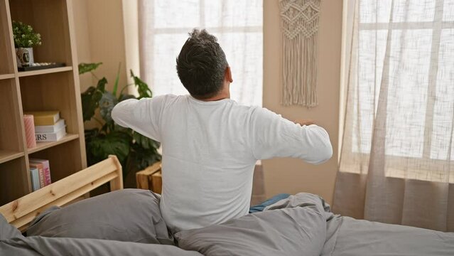 Handsome young hispanic man with grey hair stretching his arms, waking up in a cozy bed, back view in a comfortably relaxing bedroom setting