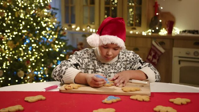 Cute smiling kid of mixed race in ornamented sweater sitting in state to art kitchen with Christmas decorations and Christmas tree with glowing garland. High quality 4k footage