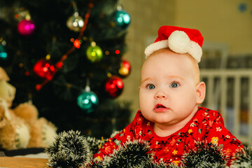 Newborn baby boy in a red Santa hat lies on the bed near the Christmas tree