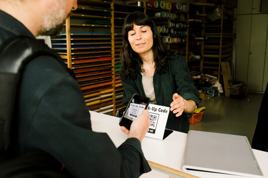 Smiling technician assisting customer scanning bar code through smart phone while doing payment at repair shop