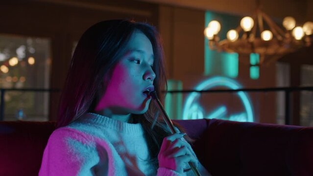 Asian young woman takes of mouthpiece to her lips, inhales and blows out puffs of white smoke at shisha cafe. Pretty adult girl smoking hookah sitting at couch at bar. Female smoker. Side view
