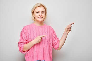 Young blond woman in pink sweater pointing aside finger, demonstrating empty space for product or...