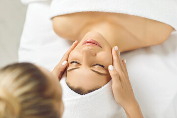 Fototapeta na wymiar Woman having spa day. Happy woman relaxing at luxury salon with white towel on head and enjoying facial massage done by professional beautician or cosmetologist. Beauty treatment, skin care concept