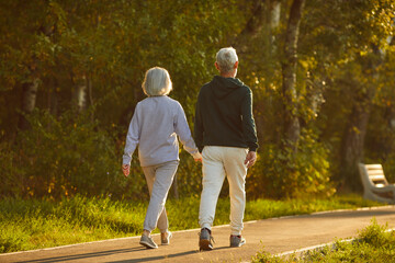 Married senior couple walking together on a sunny summer evening. From behind an old man and woman in sports clothes holding hands walking on a park walkway among green trees. Exercise, motion concept - obrazy, fototapety, plakaty