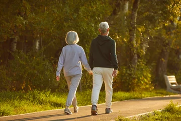 Papier Peint photo Vielles portes Married senior couple walking together on a sunny summer evening. From behind an old man and woman in sports clothes holding hands walking on a park walkway among green trees. Exercise, motion concept