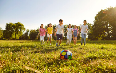 Happy children boys and girls running towards ball playing together in the park on the green grass on a summer holidays. Teenage laughing kids in casual clothes playing football in the garden. - obrazy, fototapety, plakaty