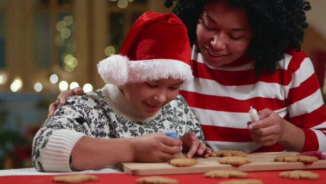 Happy black woman with kid in santa hat and sweater with ornament coloring gingerbread men in blue. Smiling caring mother helping and supporting child. High quality 4k footage