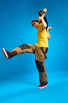 Full-length portrait of positive, smiling man dressed in rastaman, reggie style and posing with skateboard against blue studio background.