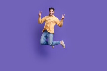 Fototapeta na wymiar Full length photo of handsome young guy jumping show v-sign wear trendy yellow garment isolated on violet color background