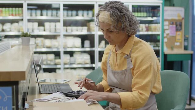 Medium shot of Caucasian female supermarket worker sitting by laptop, looking through receipts and counting on calculator while doing regular audit in morning