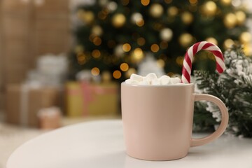 Delicious cocoa with marshmallows and candy cane in cup against blurred Christmas lights, closeup. Space for text