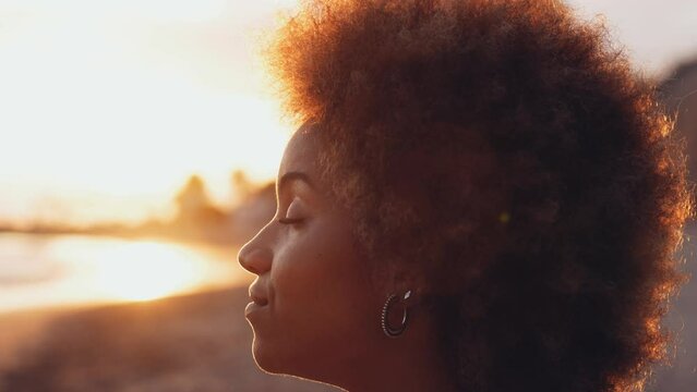 Close up portrait of one young smiling woman looking at the ocean at the beach with the sunset at her background. Beautiful casual millennial afro girl enjoying summer
