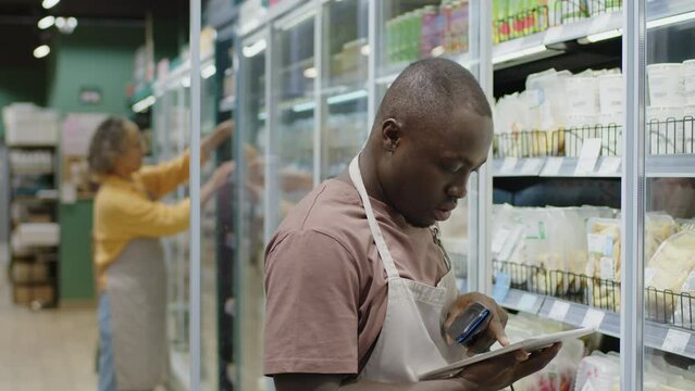 Waist up shot of African American male supermarket worker using digital tablet and scanning barcodes on ready-made products in fridge while counting inventory during morning shift