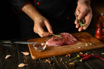 The chef adds dry aromatic rosemary to the meat steak. The process of cooking veal meat or shish kebab on the kitchen table with spices and salt