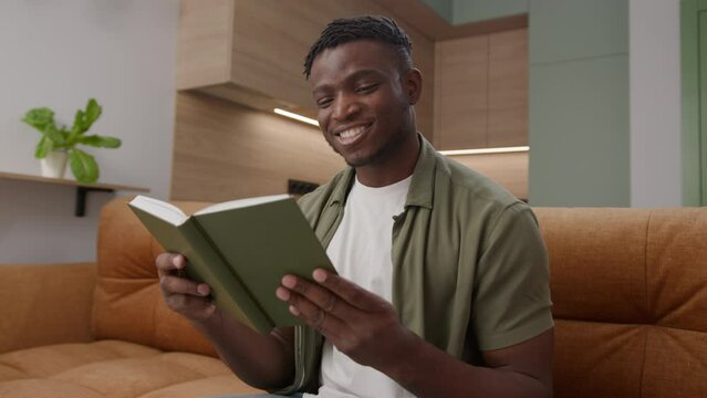 Black Man Reading Book Sitting on Sofa at Home. African American male reads paper book in living room.