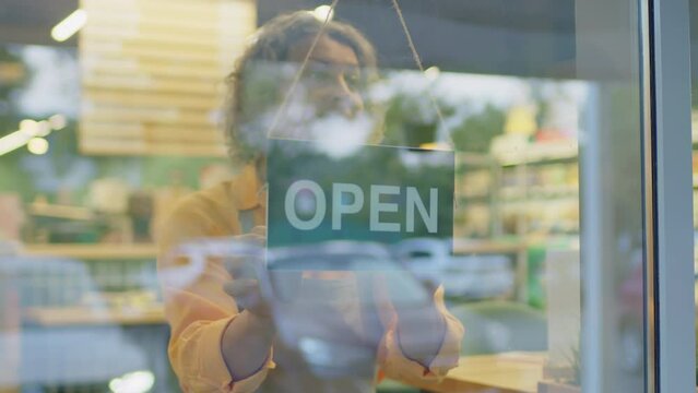 Through glass door shot of hospitable young Caucasian female food store assistant turning open sign while starting work in morning