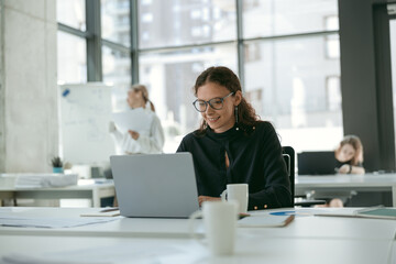 Smiling stylish businesswoman working on laptop sitting in modern office on colleagues background