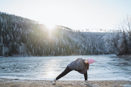 The girl practices yoga in winter in nature, asana makarasana, training in the fresh air, a frozen pond, the morning sun is shining with rays.