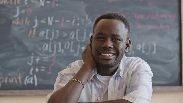 Portrait of young happy Black student sitting at desk in college classroom and smiling on camera during computer science lesson