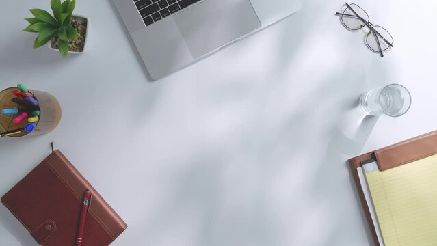 Top Down View Of A Laptop Computer Is On A White Office Desk Next To Notebook With Pens, Glasses, And A Glass Of Water. Slow Zoom In, Close Up
