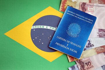 Brazilian work card and social security blue book and reais money bills on flag of Federative...