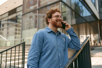 Smiling male freelancer in eyeglasses is talking phone standing on coworking building background