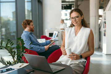 Female sales manager working on laptop and drink coffee sitting in modern coworking and looks camera