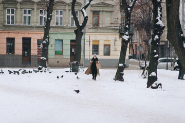 A young girl in a protective medical blue mask, a brown down jacket and a hat walks through a winter park with a bag in her hands and two glasses of coffee. Coronavirus Covid-19 pandemic. Ukraine Lviv