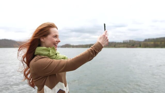 red-haired woman stands by lake feeling wind in hair holding smartphone