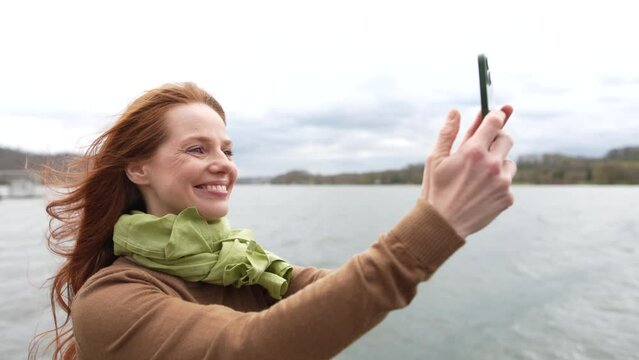 Young redhead takes photo on windy autumn day standing by lake shore