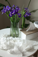 Concept of seasonal allergy to flower. Bouquet of purple iris flowers in a glass vase on table and many used napkins at home interior. Allergic rhinitis symptom of odor pollen flowering