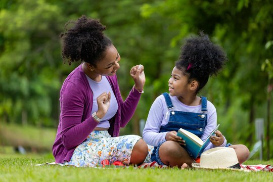 African American mother is teaching her young daughter to read using digital tablet while having a summer picnic in public park for education and happiness