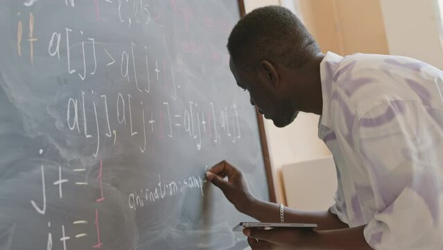 Young African American college student holding digital tablet and writing computer algorithm on chalkboard in classroom