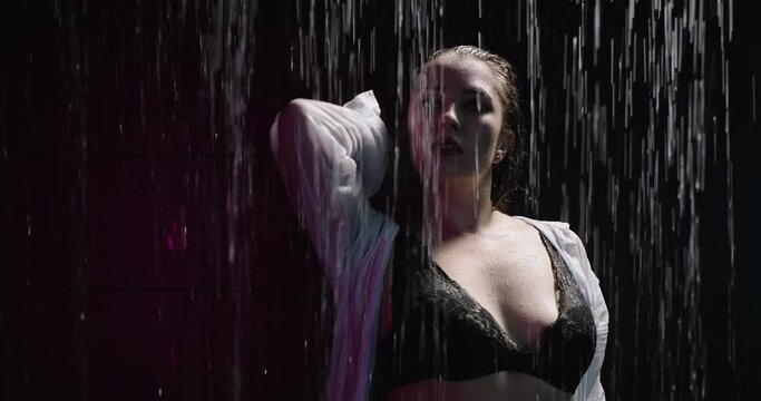 Close-up of wet woman standing in the shower and looking into the camera, water drops flow down the body. Sexy woman in black lingerie and white man shirt sensual moves. Slow motion