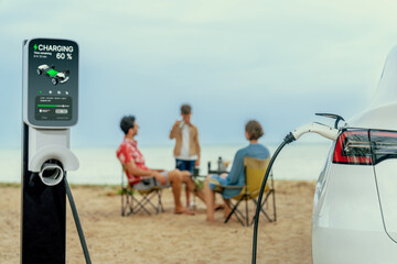Alternative family vacation trip traveling by the beach with electric car recharging battery from EV charging station with blurred family enjoying the seascape background. Perpetual - obrazy, fototapety, plakaty