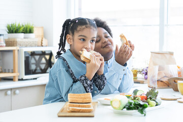 African little child girl eating homemade sandwiches in hands, standing in kitchen at home with brother, prepare food for breakfast. Siblings, kids make sandwich. Selective focus on girl - obrazy, fototapety, plakaty