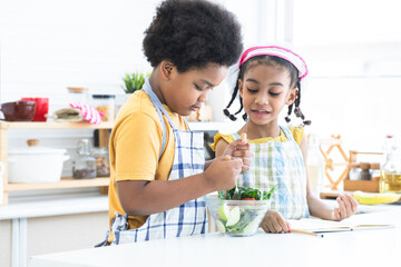 African siblings, boy in apron cooking, making mixing salad in bowl with little girl wear chef hat, holding pencil take note recipes on book in kitchen at home. Children education and healthy food
