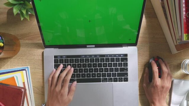 Top Down View Of Man'S Hands Typing On Mock Up Green Screen Chromakey Laptop And Using Wireless Mouse On A Wooden Desk With Books, Headphones, Pens, Glasses, And A Glass Of Water. Zoom Out, Close Up
